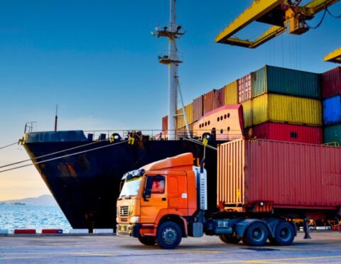 Truck carrying forty-foot container leaving port terminal with ship and quay crane on the background.